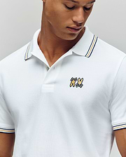 A man wears the Psycho Bunny MENS ARCHIVE NEON BUNNY POLO (MT0100018), a white 100% Pima cotton regular fit polo with dark-striped collar and sleeves, featuring embroidered Bunny and cartoon animal on the left chest. He looks downward. (3)