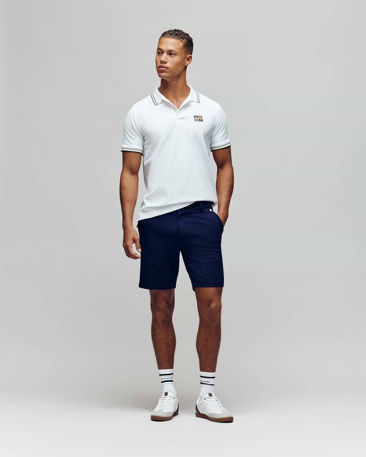 A man stands against a plain background wearing the Psycho Bunny MENS ARCHIVE NEON BUNNY POLO - MT0100018 in white, paired with navy shorts, striped white socks, and white sneakers. He has one hand in his pocket and looks slightly to the side. (2)