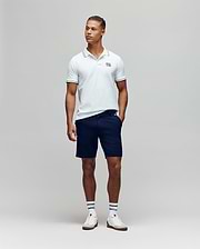 A man stands against a plain background wearing the Psycho Bunny MENS ARCHIVE NEON BUNNY POLO - MT0100018 in white, paired with navy shorts, striped white socks, and white sneakers. He has one hand in his pocket and looks slightly to the side. (2)