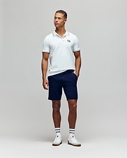 A man stands against a plain background wearing the Psycho Bunny MENS ARCHIVE NEON BUNNY POLO - MT0100018 in white, paired with navy shorts, striped white socks, and white sneakers. He has one hand in his pocket and looks slightly to the side. (2)
