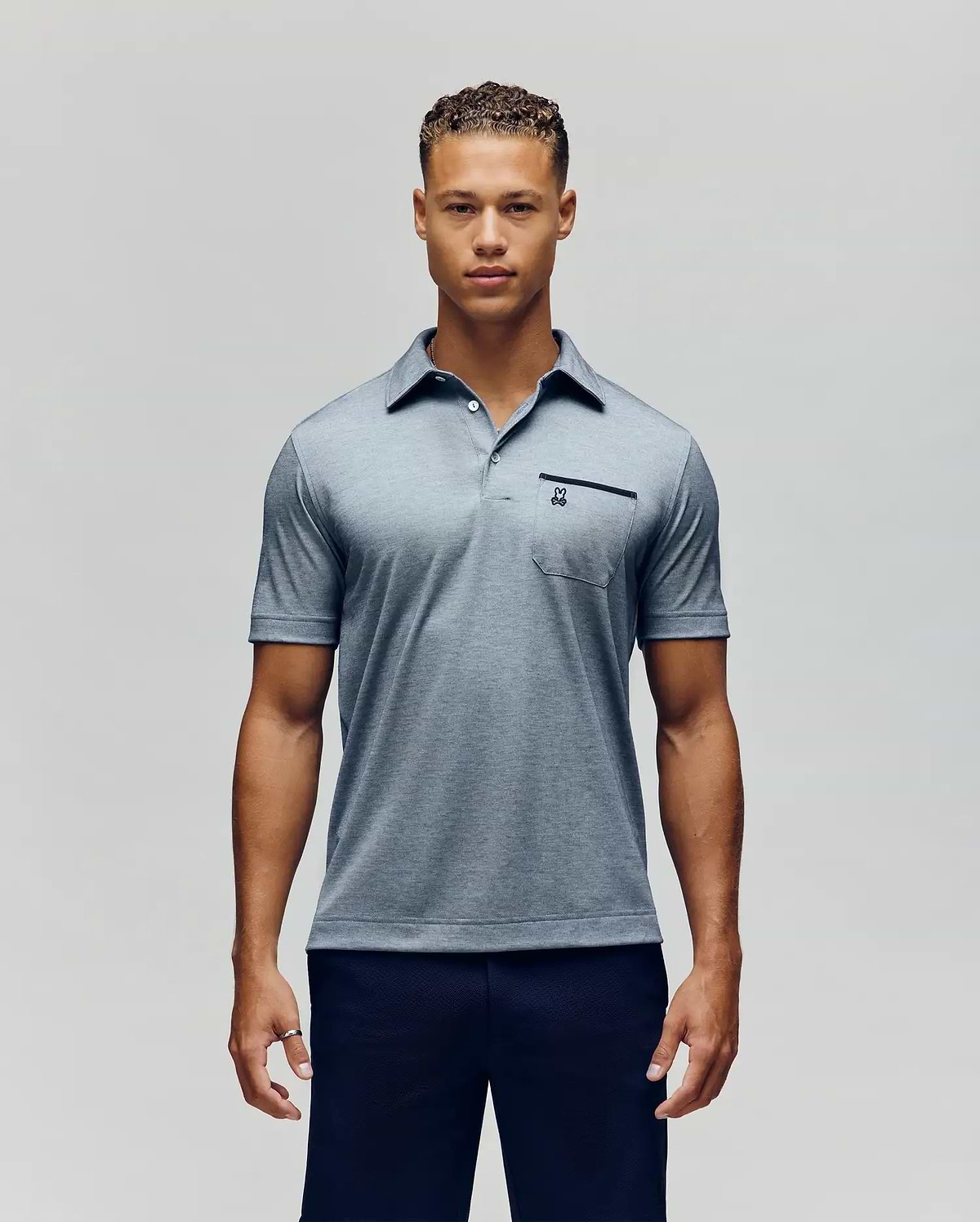 A man poses against a plain background, wearing the Psycho Bunny MENS HADLEY PERFORMANCE PIQUE POLO - MT0100021 in light blue with a buttoned collar and small chest pocket, paired with dark pants, looking at the camera with a neutral expression. (1)