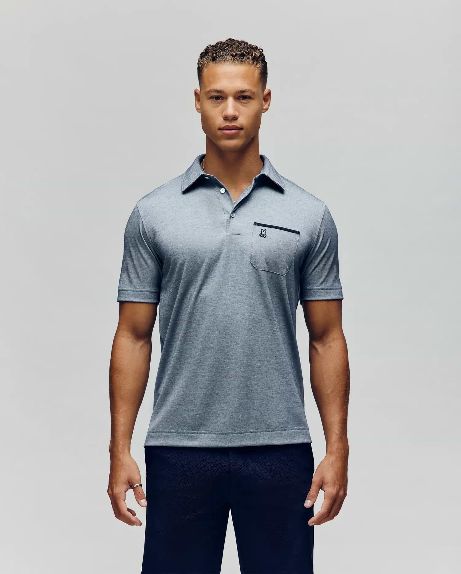 A man poses against a plain background, wearing the Psycho Bunny MENS HADLEY PERFORMANCE PIQUE POLO - MT0100021 in light blue with a buttoned collar and small chest pocket, paired with dark pants, looking at the camera with a neutral expression. (1)