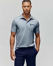 A young man with short curly hair wears the Psycho Bunny MENS HADLEY PERFORMANCE PIQUE POLO in light gray and dark navy pants, standing against a plain light gray background. (2)