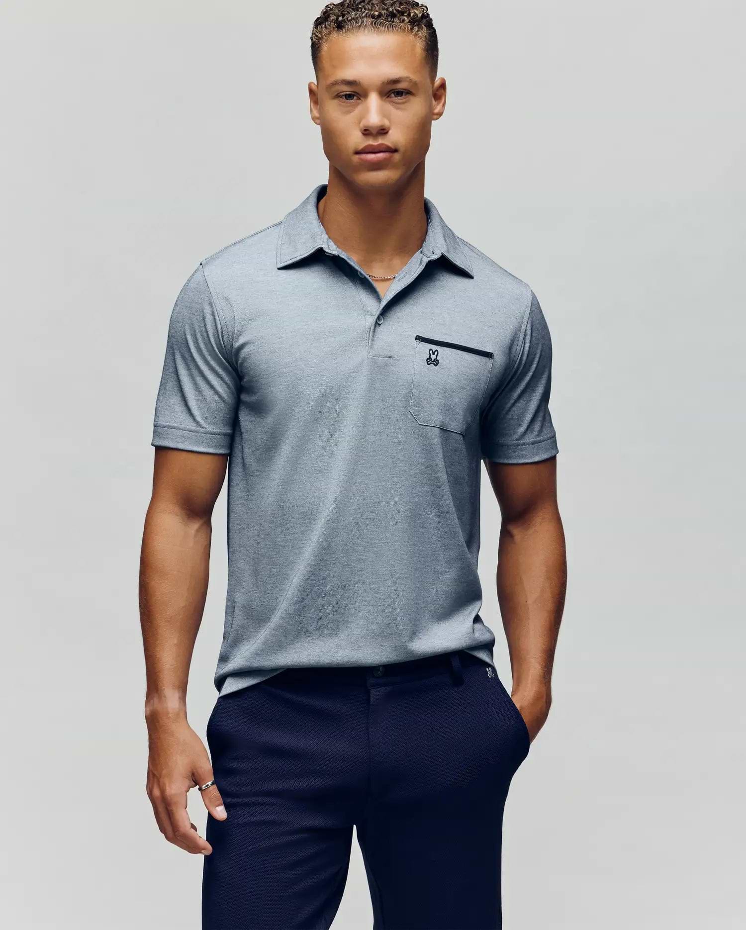 A young man with short curly hair wears the Psycho Bunny MENS HADLEY PERFORMANCE PIQUE POLO in light gray and dark navy pants, standing against a plain light gray background. (2)