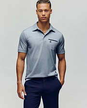 A young man with short curly hair wears the Psycho Bunny MENS HADLEY PERFORMANCE PIQUE POLO in light gray and dark navy pants, standing against a plain light gray background. (2)