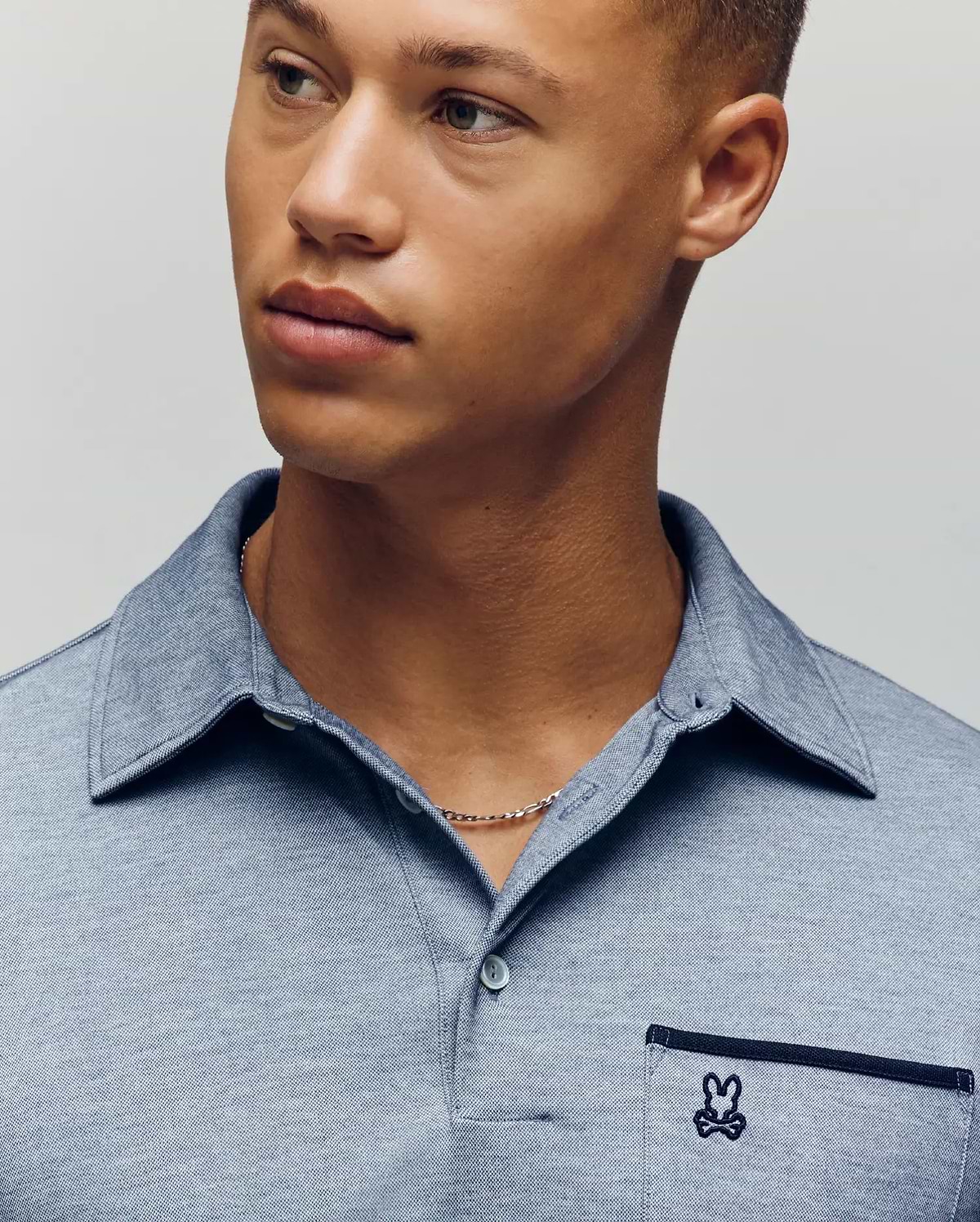 A young man with light brown skin and short hair wears the Psycho Bunny MENS HADLEY PERFORMANCE PIQUE POLO (MT0100021), a light blue collared shirt featuring the brand’s signature small embroidered bunny logo on the pocket. (3)