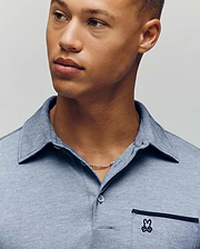 A young man with light brown skin and short hair wears the Psycho Bunny MENS HADLEY PERFORMANCE PIQUE POLO (MT0100021), a light blue collared shirt featuring the brand’s signature small embroidered bunny logo on the pocket. (3)