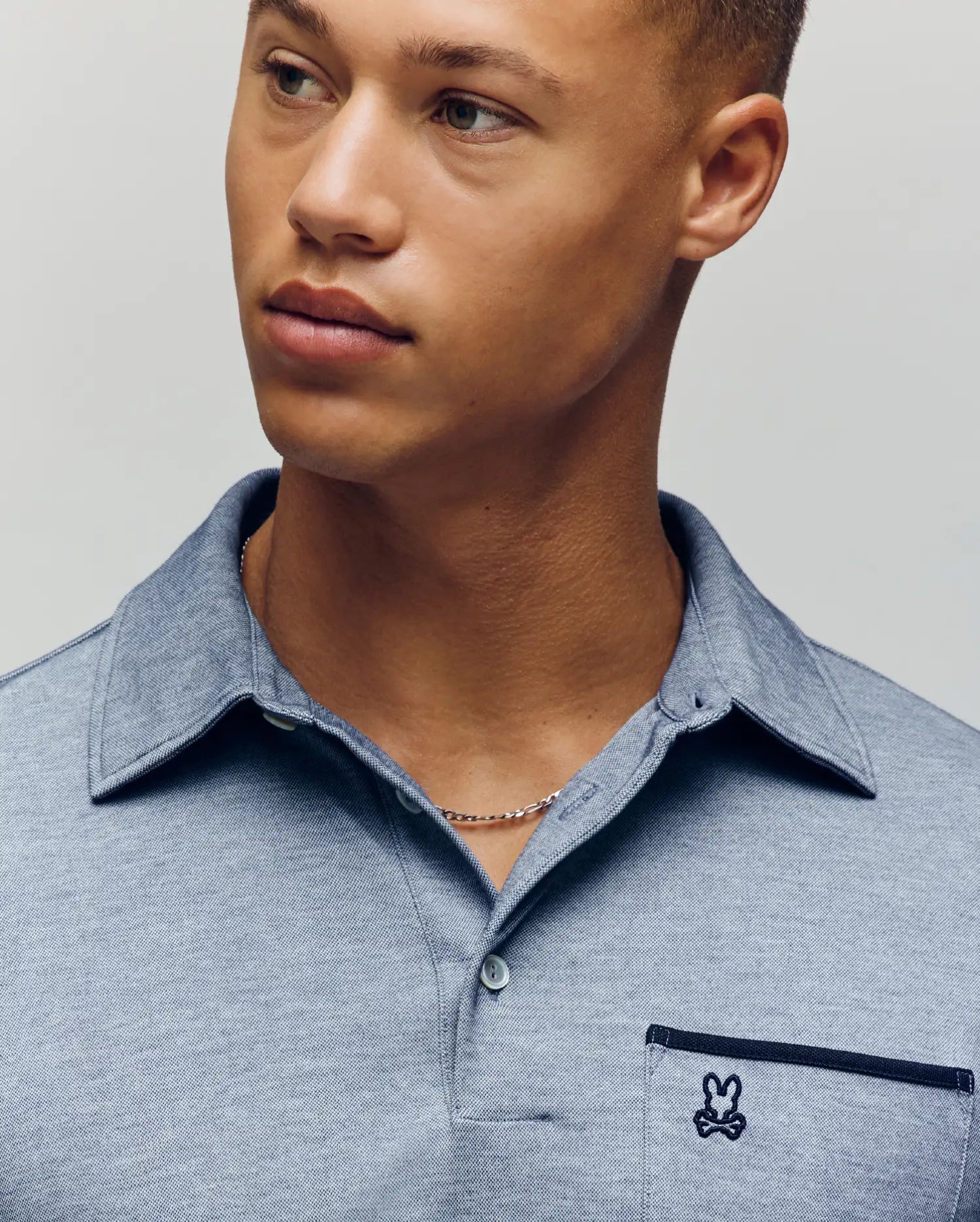 A young man with light brown skin and short hair wears the Psycho Bunny MENS HADLEY PERFORMANCE PIQUE POLO (MT0100021), a light blue collared shirt featuring the brand’s signature small embroidered bunny logo on the pocket. (3)