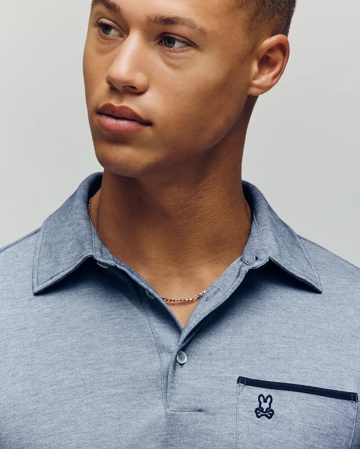 A young man with light brown skin and short hair wears the Psycho Bunny MENS HADLEY PERFORMANCE PIQUE POLO (MT0100021), a light blue collared shirt featuring the brand’s signature small embroidered bunny logo on the pocket. (3)