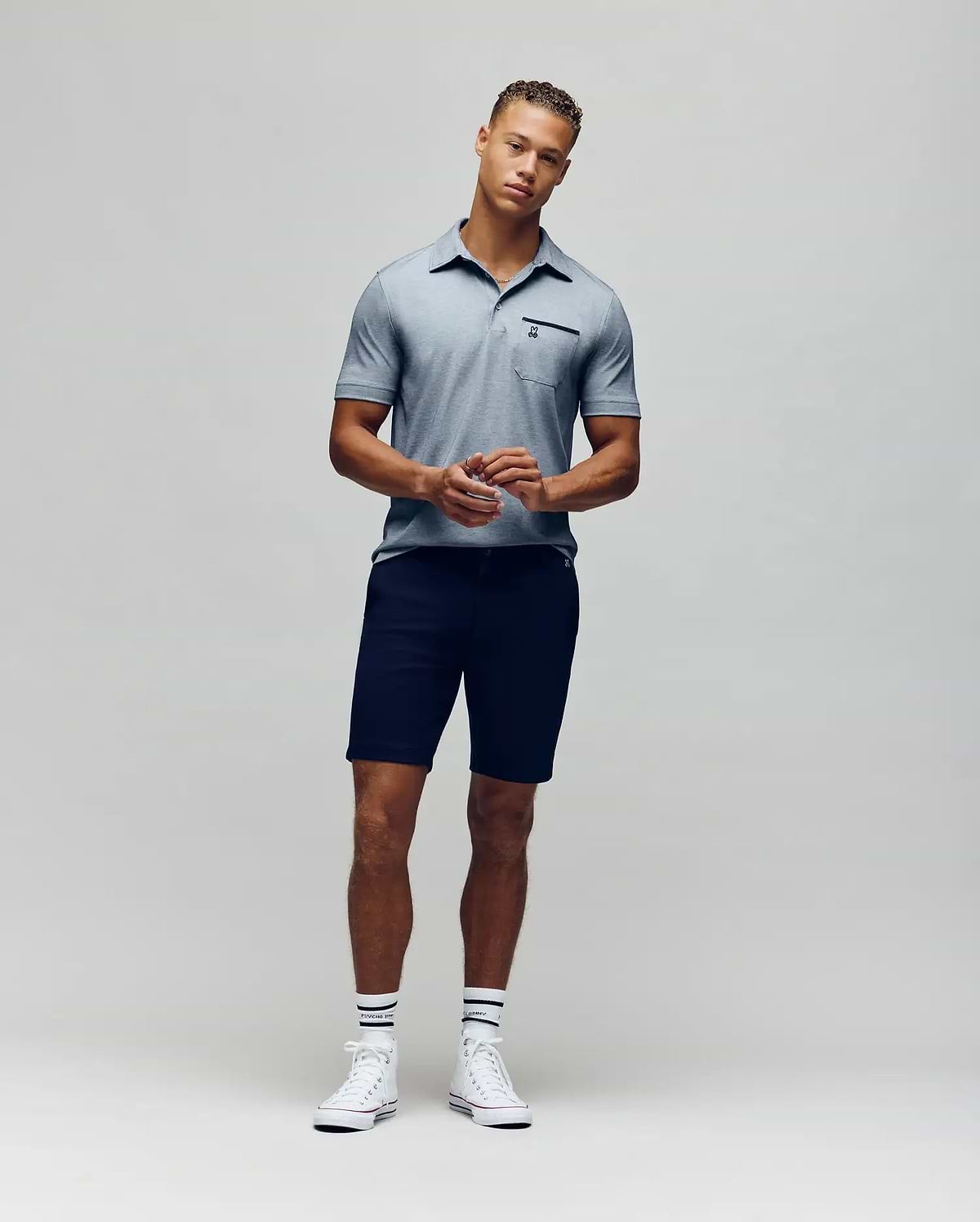 A man stands against a plain background wearing the Psycho Bunny MENS HADLEY PERFORMANCE PIQUE POLO (MT0100021), dark shorts, striped white socks, and white sneakers. He faces the camera with his hands clasped in front of him. (4)