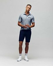 A man stands against a plain background wearing the Psycho Bunny MENS HADLEY PERFORMANCE PIQUE POLO (MT0100021), dark shorts, striped white socks, and white sneakers. He faces the camera with his hands clasped in front of him. (4)