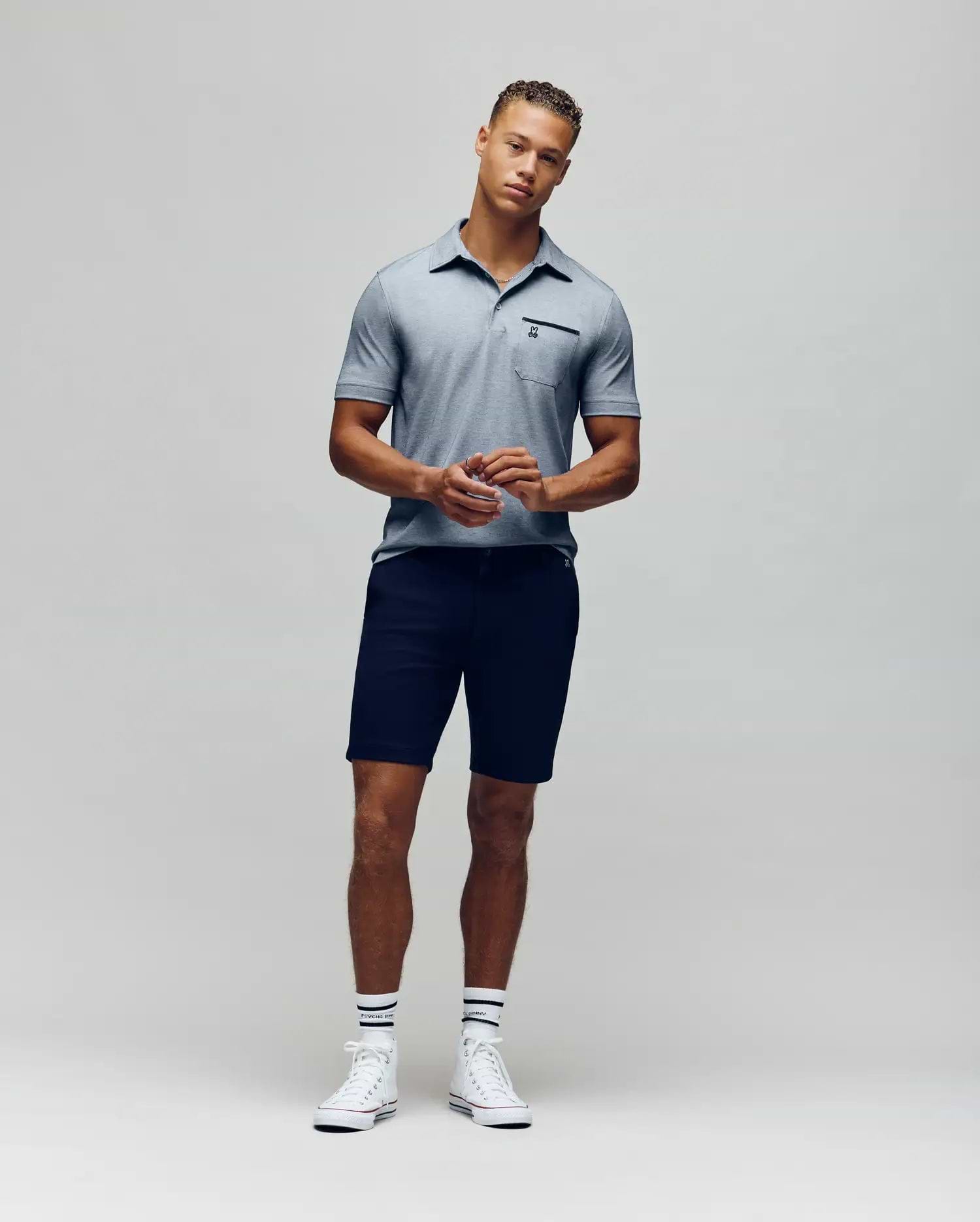 A man stands against a plain background wearing the Psycho Bunny MENS HADLEY PERFORMANCE PIQUE POLO (MT0100021), dark shorts, striped white socks, and white sneakers. He faces the camera with his hands clasped in front of him. (4)
