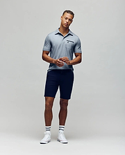 A man stands against a plain background wearing the Psycho Bunny MENS HADLEY PERFORMANCE PIQUE POLO (MT0100021), dark shorts, striped white socks, and white sneakers. He faces the camera with his hands clasped in front of him. (4)