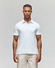 A man with short curly hair stands facing forward in the Psycho Bunny MENS HADLEY PERFORMANCE PIQUE POLO (MT0100021), a white short-sleeve shirt with a chest pocket, paired with light-colored shorts against a plain background. (1)
