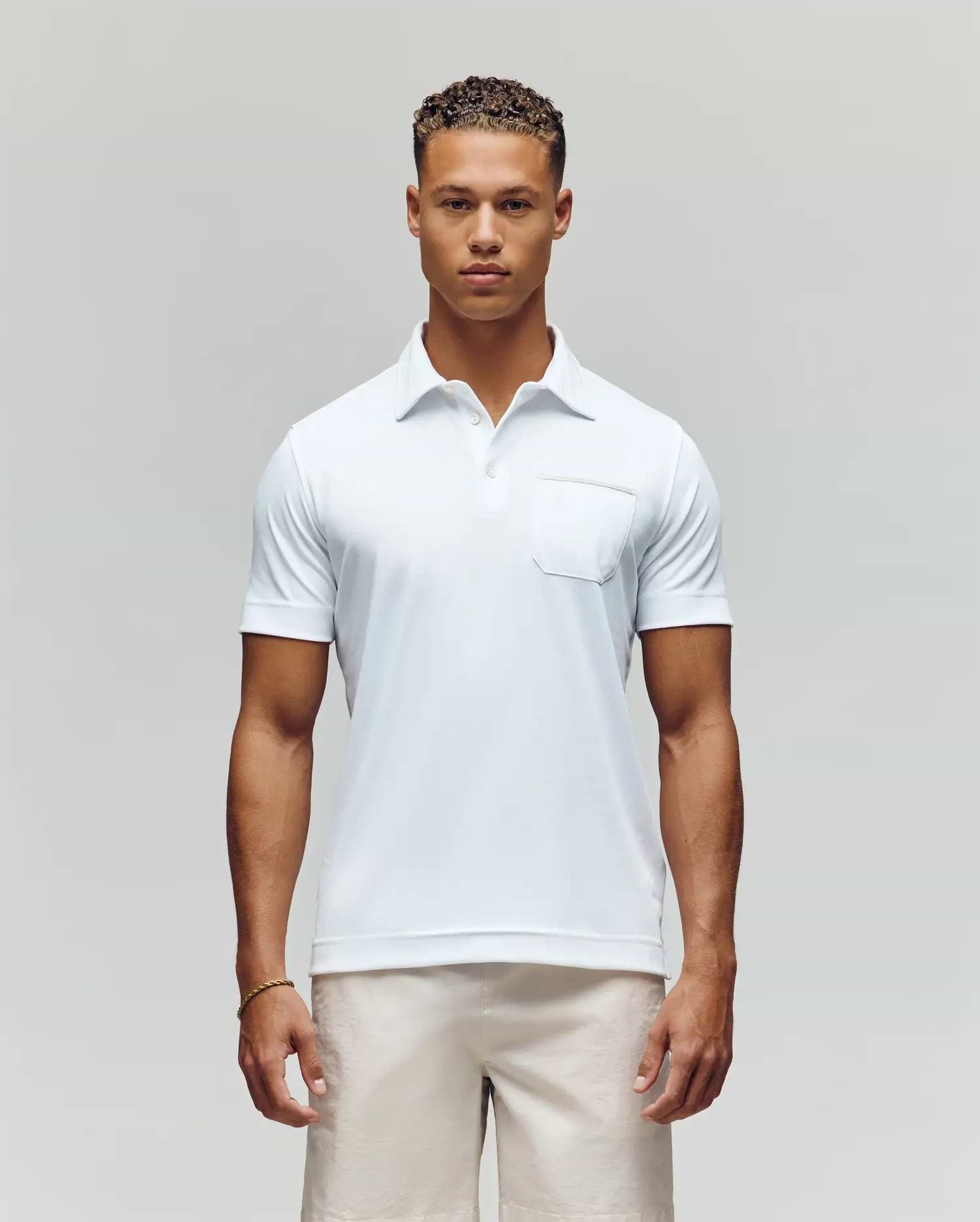 A man with short curly hair stands facing forward in the Psycho Bunny MENS HADLEY PERFORMANCE PIQUE POLO (MT0100021), a white short-sleeve shirt with a chest pocket, paired with light-colored shorts against a plain background. (1)