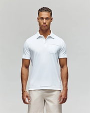 A man with short curly hair stands facing forward in the Psycho Bunny MENS HADLEY PERFORMANCE PIQUE POLO (MT0100021), a white short-sleeve shirt with a chest pocket, paired with light-colored shorts against a plain background. (1)