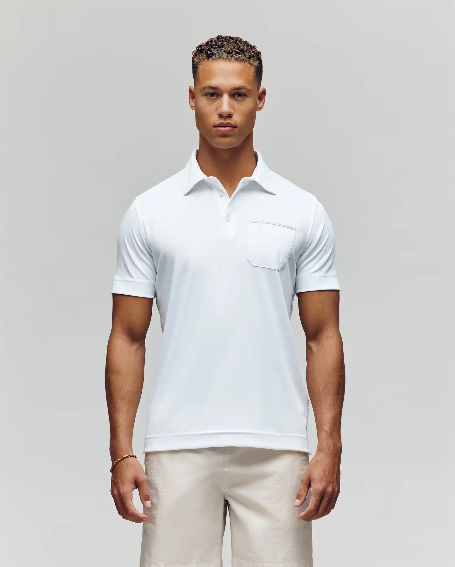 A man with short curly hair stands facing forward in the Psycho Bunny MENS HADLEY PERFORMANCE PIQUE POLO (MT0100021), a white short-sleeve shirt with a chest pocket, paired with light-colored shorts against a plain background. (1)