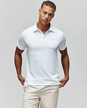 A man with short curly hair models the Psycho Bunny MENS HADLEY PERFORMANCE PIQUE POLO - MT0100021, a white short-sleeve polo featuring a chest pocket, paired with light shorts, standing against a plain light background with hands in his pockets. (2)