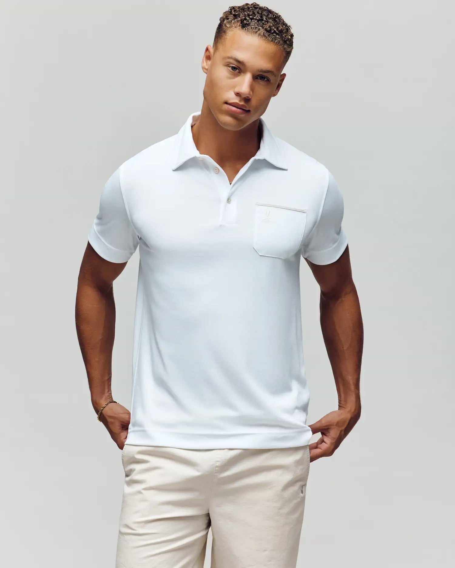 A man with short curly hair models the Psycho Bunny MENS HADLEY PERFORMANCE PIQUE POLO - MT0100021, a white short-sleeve polo featuring a chest pocket, paired with light shorts, standing against a plain light background with hands in his pockets. (2)