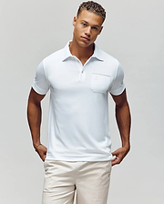 A man with short curly hair models the Psycho Bunny MENS HADLEY PERFORMANCE PIQUE POLO - MT0100021, a white short-sleeve polo featuring a chest pocket, paired with light shorts, standing against a plain light background with hands in his pockets. (2)