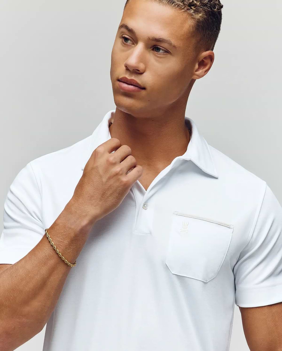 A young man in a Psycho Bunny MENS HADLEY PERFORMANCE PIQUE POLO (MT0100021) and a gold bracelet looks thoughtfully to the side, touching his collar with one hand against a plain, light background. (3)