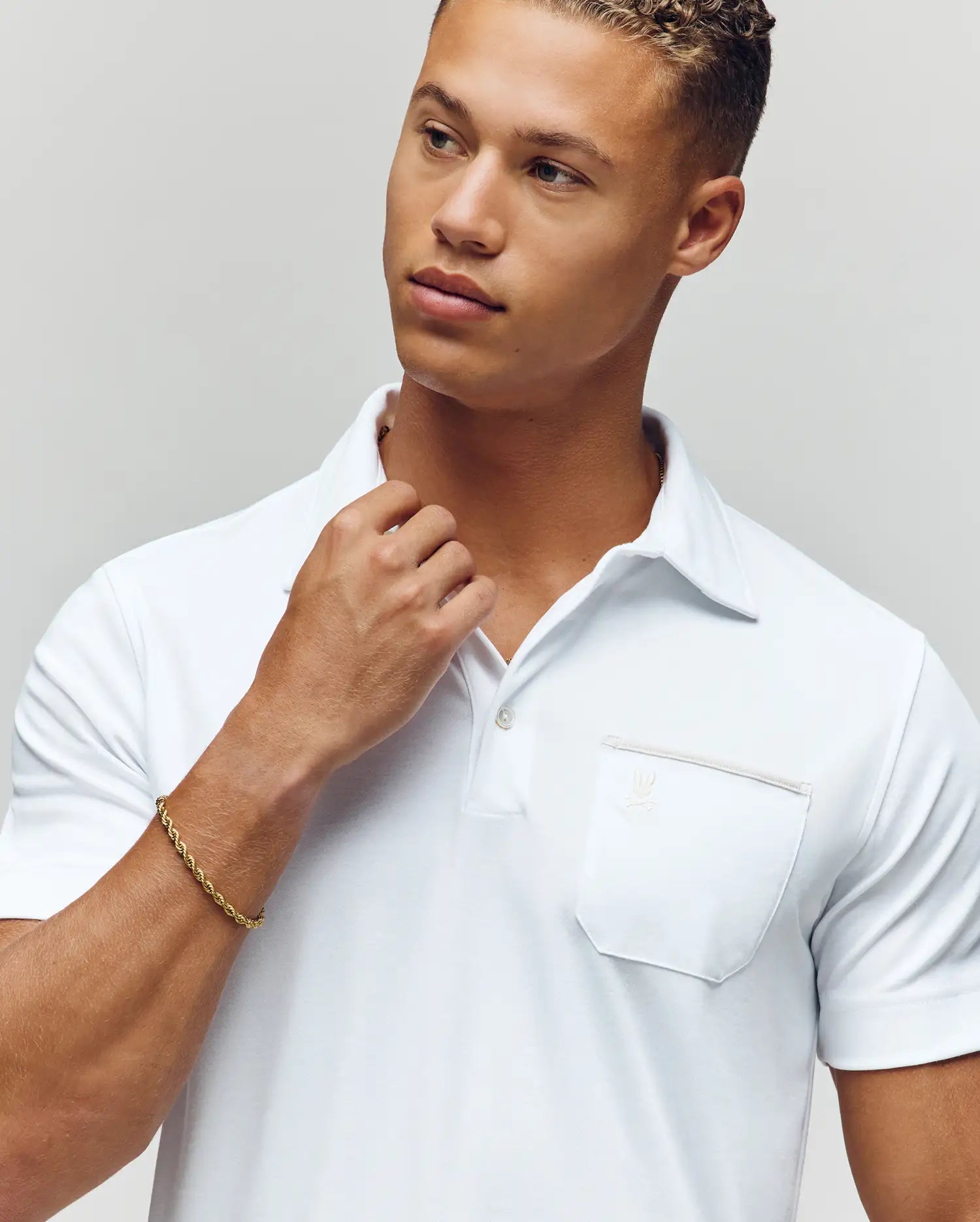 A young man in a Psycho Bunny MENS HADLEY PERFORMANCE PIQUE POLO (MT0100021) and a gold bracelet looks thoughtfully to the side, touching his collar with one hand against a plain, light background. (3)