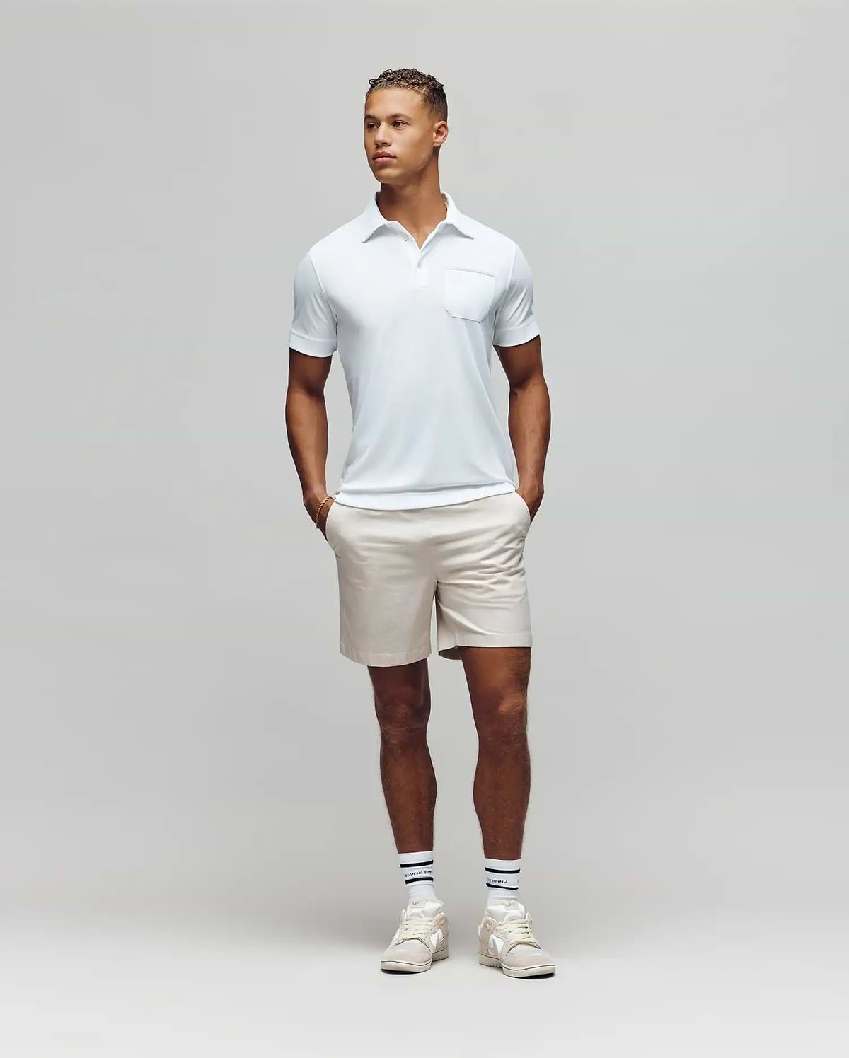 A man stands against a plain light gray background, wearing the Psycho Bunny MENS HADLEY PERFORMANCE PIQUE POLO - MT0100021 in light blue, paired with beige shorts and white athletic shoes, hands in pockets and looking slightly to his left. (4)