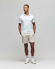 A man stands against a plain light gray background, wearing the Psycho Bunny MENS HADLEY PERFORMANCE PIQUE POLO - MT0100021 in light blue, paired with beige shorts and white athletic shoes, hands in pockets and looking slightly to his left. (4)