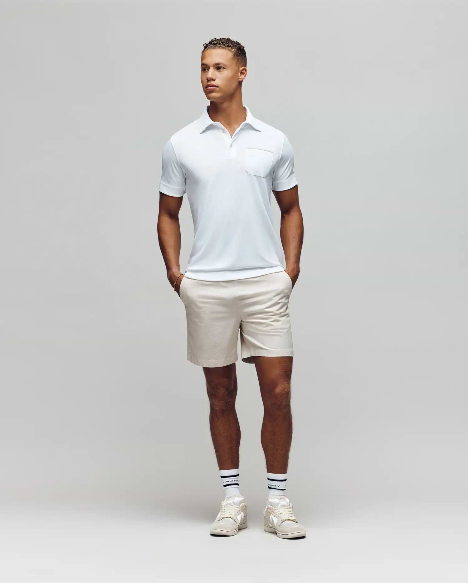 A man stands against a plain light gray background, wearing the Psycho Bunny MENS HADLEY PERFORMANCE PIQUE POLO - MT0100021 in light blue, paired with beige shorts and white athletic shoes, hands in pockets and looking slightly to his left. (4)