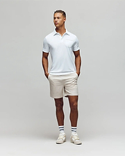 A man stands against a plain light gray background, wearing the Psycho Bunny MENS HADLEY PERFORMANCE PIQUE POLO - MT0100021 in light blue, paired with beige shorts and white athletic shoes, hands in pockets and looking slightly to his left. (4)