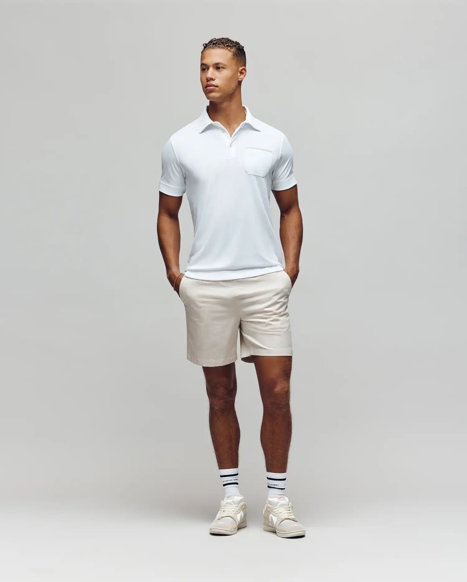 A man stands against a plain light gray background, wearing the Psycho Bunny MENS HADLEY PERFORMANCE PIQUE POLO - MT0100021 in light blue, paired with beige shorts and white athletic shoes, hands in pockets and looking slightly to his left. (4)