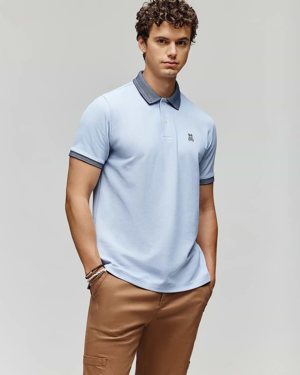 A young man with curly hair wears the Psycho Bunny MENS MURPHY PIQUE POLO SHIRT - MT0100026 in light blue with a dark collar, embroidered Bunny logo, and tan pants, posing with hands in pockets against a plain light gray background. (1)
