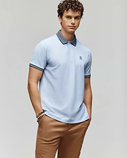 A young man with curly hair wears the Psycho Bunny MENS MURPHY PIQUE POLO SHIRT - MT0100026 in light blue with a dark collar, embroidered Bunny logo, and tan pants, posing with hands in pockets against a plain light gray background. (1)