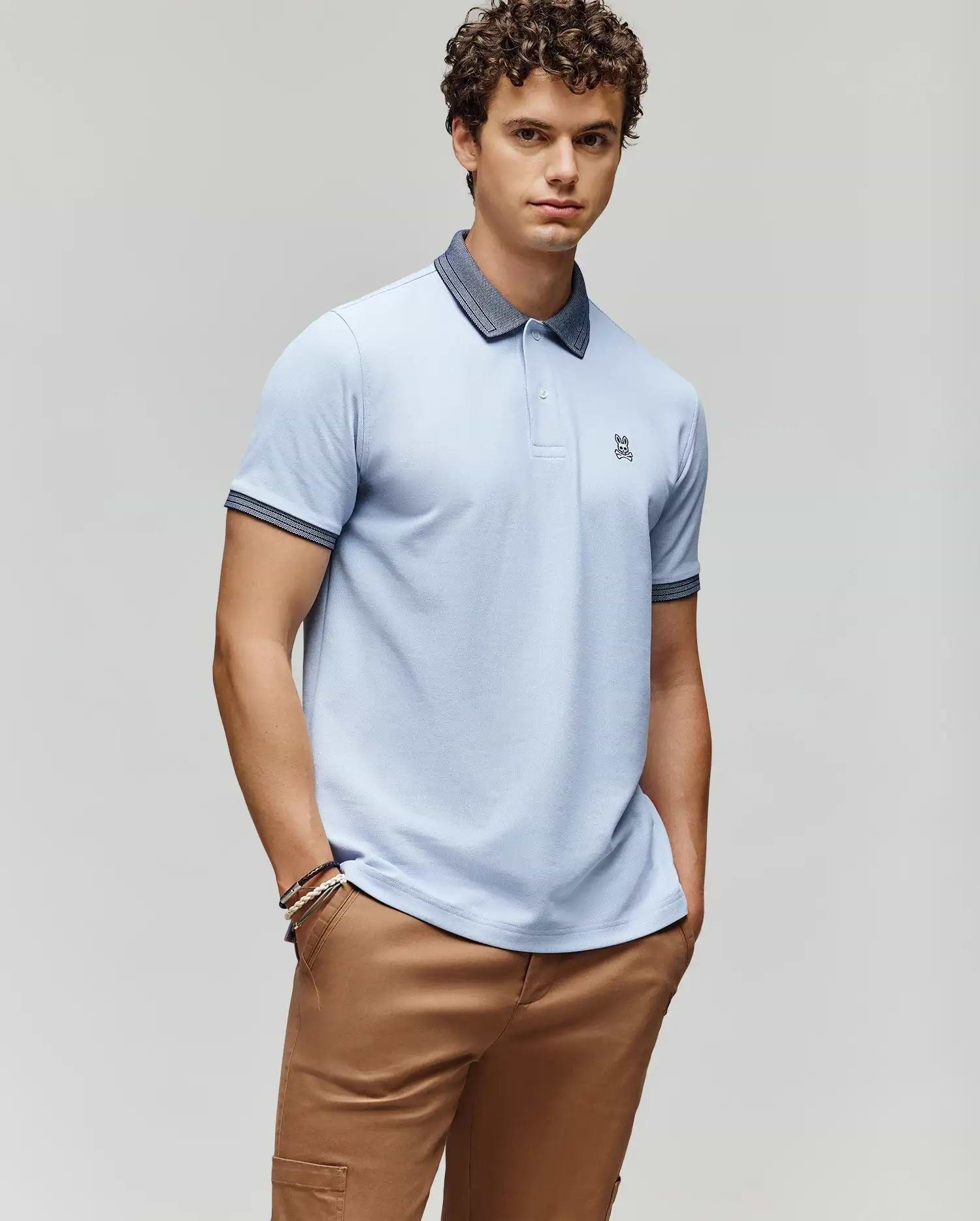 A young man with curly hair wears the Psycho Bunny MENS MURPHY PIQUE POLO SHIRT - MT0100026 in light blue with a dark collar, embroidered Bunny logo, and tan pants, posing with hands in pockets against a plain light gray background. (1)