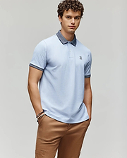 A young man with curly hair wears the Psycho Bunny MENS MURPHY PIQUE POLO SHIRT - MT0100026 in light blue with a dark collar, embroidered Bunny logo, and tan pants, posing with hands in pockets against a plain light gray background. (1)