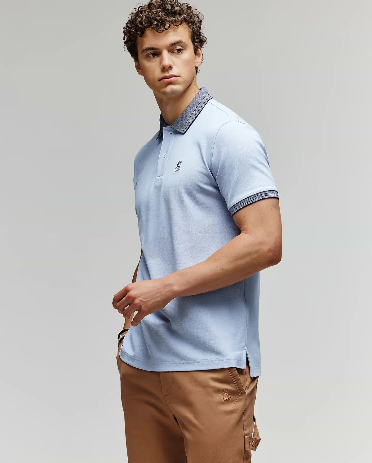A man with curly hair wears a light blue Psycho Bunny MENS MURPHY PIQUE POLO SHIRT (MT0100026) with a dark collar and tan pants, standing against a plain background, looking to the side with one hand on his hip. (3)