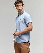 A man with curly hair wears a light blue Psycho Bunny MENS MURPHY PIQUE POLO SHIRT (MT0100026) with a dark collar and tan pants, standing against a plain background, looking to the side with one hand on his hip. (3)