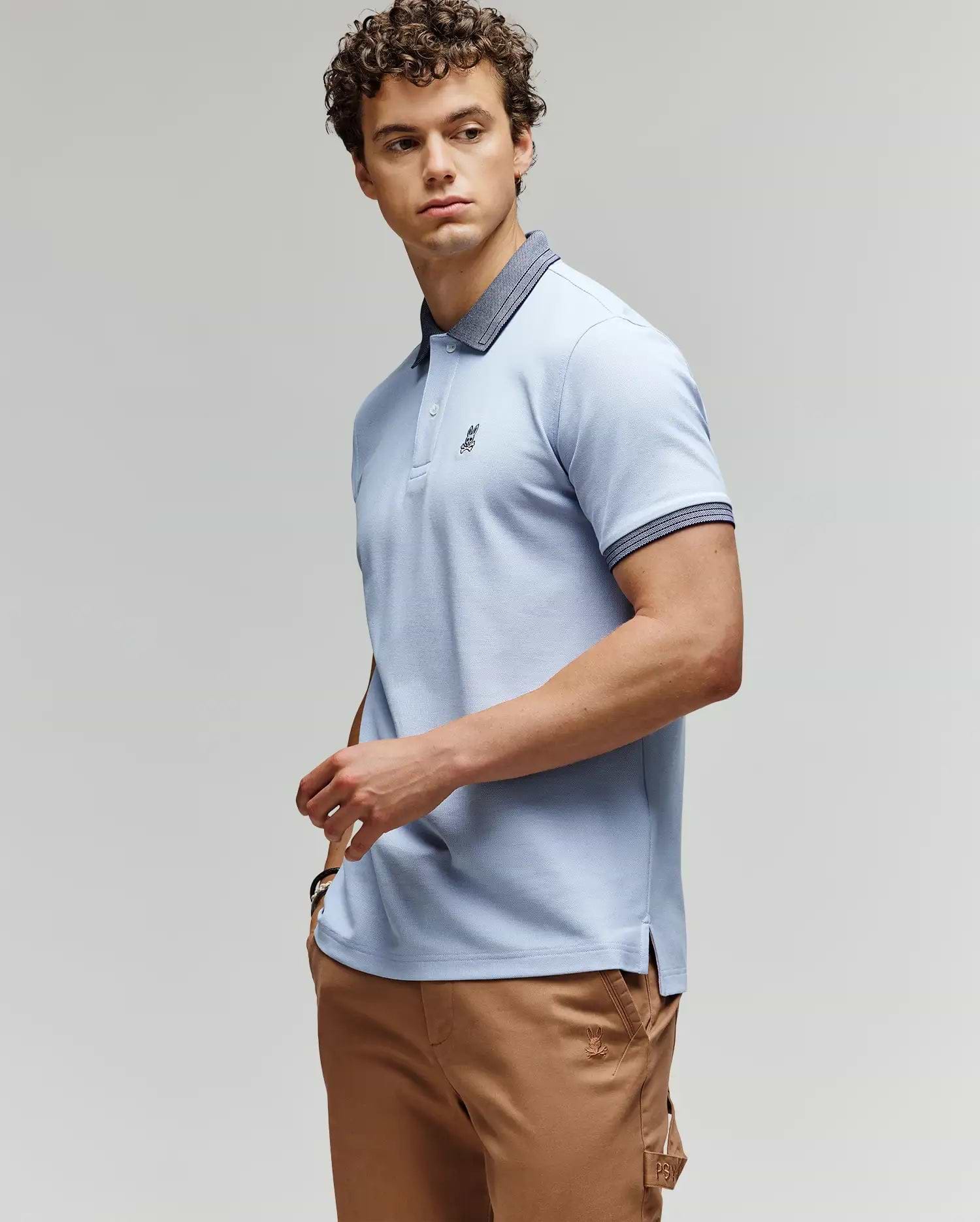A man with curly hair wears a light blue Psycho Bunny MENS MURPHY PIQUE POLO SHIRT (MT0100026) with a dark collar and tan pants, standing against a plain background, looking to the side with one hand on his hip. (3)