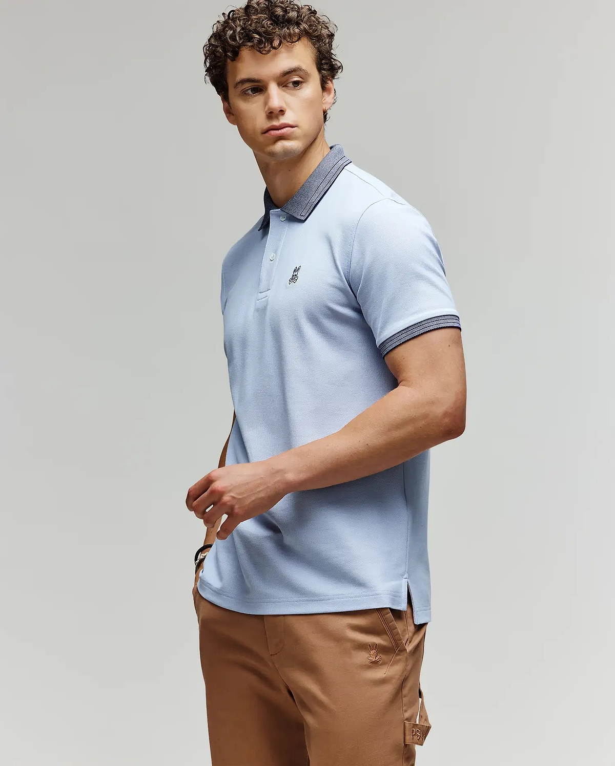 A man with curly hair wears a light blue Psycho Bunny MENS MURPHY PIQUE POLO SHIRT (MT0100026) with a dark collar and tan pants, standing against a plain background, looking to the side with one hand on his hip. (3)