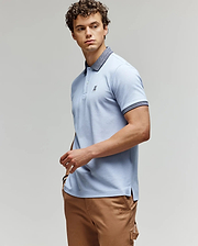 A man with curly hair wears a light blue Psycho Bunny MENS MURPHY PIQUE POLO SHIRT (MT0100026) with a dark collar and tan pants, standing against a plain background, looking to the side with one hand on his hip. (3)