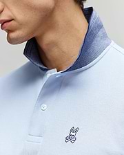 A close-up of a person wearing the Psycho Bunny MENS MURPHY PIQUE POLO SHIRT - MT0100026, a light blue Pima cotton polo with a dark blue inner collar and an embroidered Bunny logo and crossbones on the chest. The face is partially visible. (2)