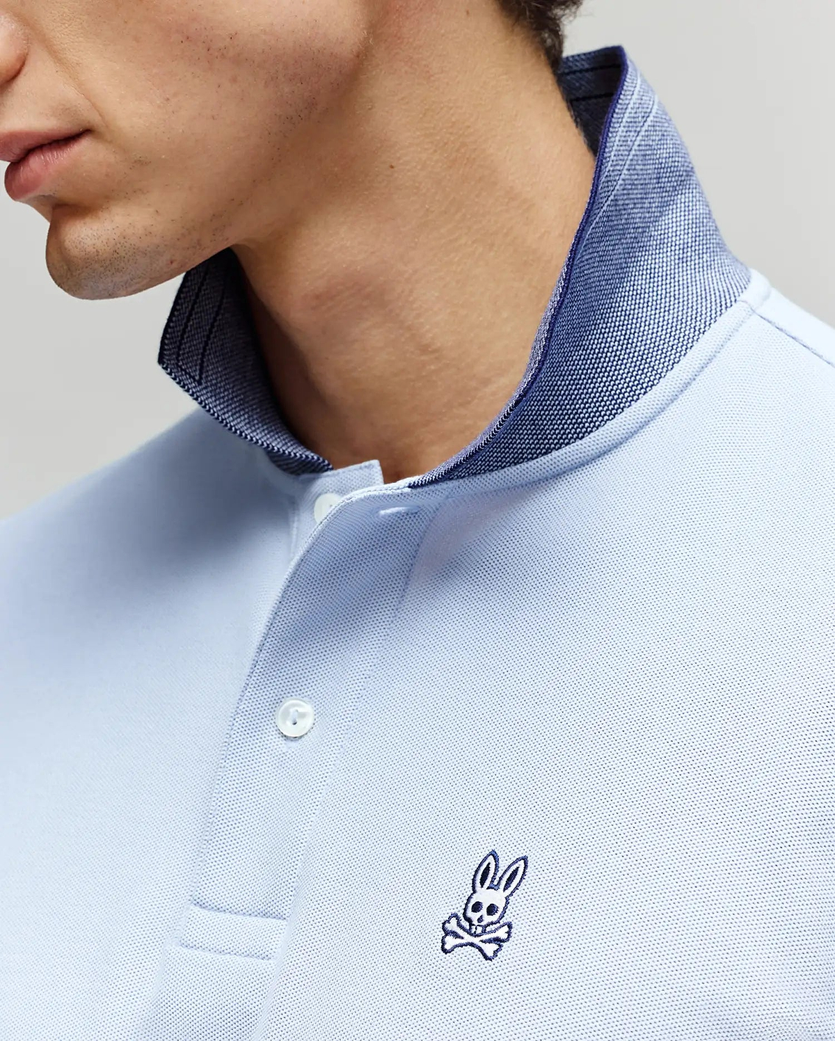 A close-up of a person wearing the Psycho Bunny MENS MURPHY PIQUE POLO SHIRT - MT0100026, a light blue Pima cotton polo with a dark blue inner collar and an embroidered Bunny logo and crossbones on the chest. The face is partially visible. (2)