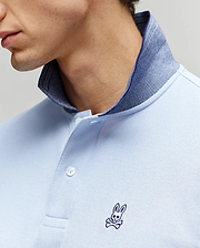 A close-up of a person wearing the Psycho Bunny MENS MURPHY PIQUE POLO SHIRT - MT0100026, a light blue Pima cotton polo with a dark blue inner collar and an embroidered Bunny logo and crossbones on the chest. The face is partially visible. (2)
