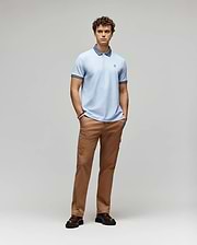 A young man stands with hands in pockets, wearing the Psycho Bunny MENS MURPHY PIQUE POLO SHIRT (MT0100026) in light blue with an embroidered Bunny logo, tan pants, and dark brown shoes, against a plain light gray background. (4)