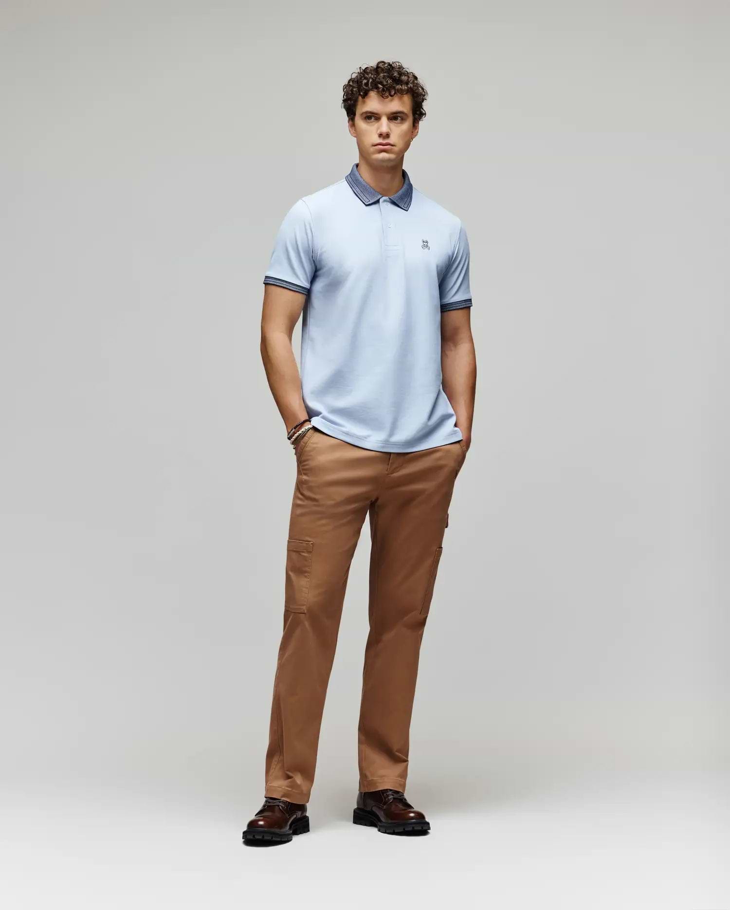 A young man stands with hands in pockets, wearing the Psycho Bunny MENS MURPHY PIQUE POLO SHIRT (MT0100026) in light blue with an embroidered Bunny logo, tan pants, and dark brown shoes, against a plain light gray background. (4)