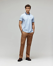 A young man stands with hands in pockets, wearing the Psycho Bunny MENS MURPHY PIQUE POLO SHIRT (MT0100026) in light blue with an embroidered Bunny logo, tan pants, and dark brown shoes, against a plain light gray background. (4)