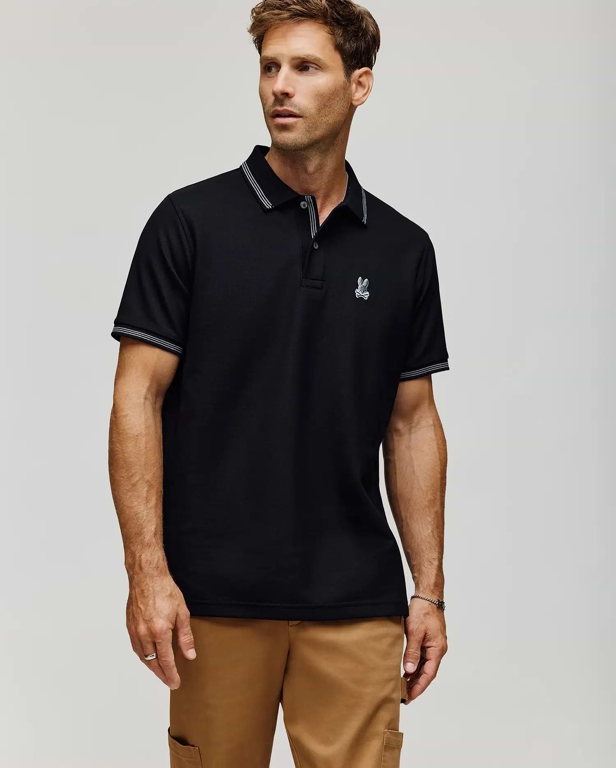 A man wears the Psycho Bunny MENS HART PIQUE POLO SHIRT - MT0100032 in black, featuring white trim on the collar and sleeves and a small embroidered rabbit logo on the left chest, paired with tan pants against a plain light background. (1)