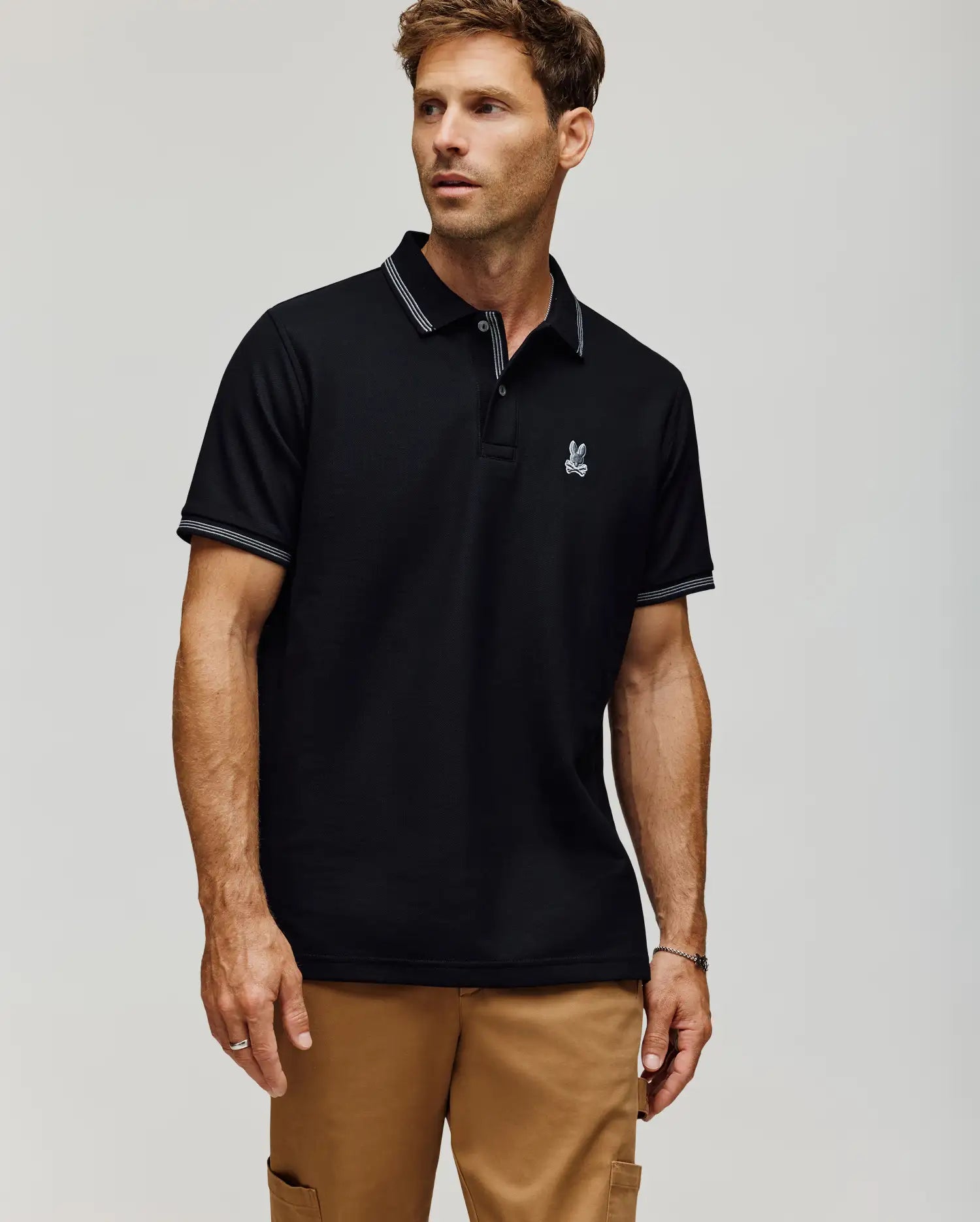 A man wears the Psycho Bunny MENS HART PIQUE POLO SHIRT - MT0100032 in black, featuring white trim on the collar and sleeves and a small embroidered rabbit logo on the left chest, paired with tan pants against a plain light background. (1)