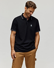 A man in the Psycho Bunny MENS HART PIQUE POLO SHIRT (MT0100032), featuring soft Pima cotton and an embroidered white bunny logo, pairs it with tan pants and stands smiling with hands in pockets against a light background. (3)