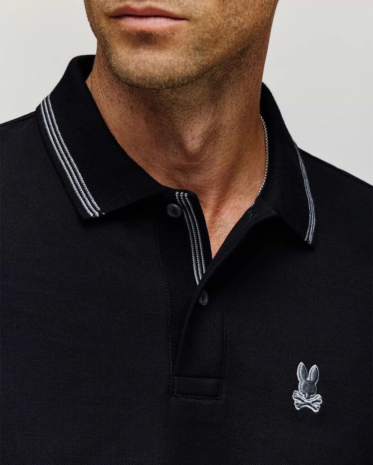 A man wears the Psycho Bunny MENS HART PIQUE POLO SHIRT (MT0100032), a black designer polo with white trim and a small bunny with crossbones logo on the chest; image shows only his lower face, neck, and upper torso. (2)